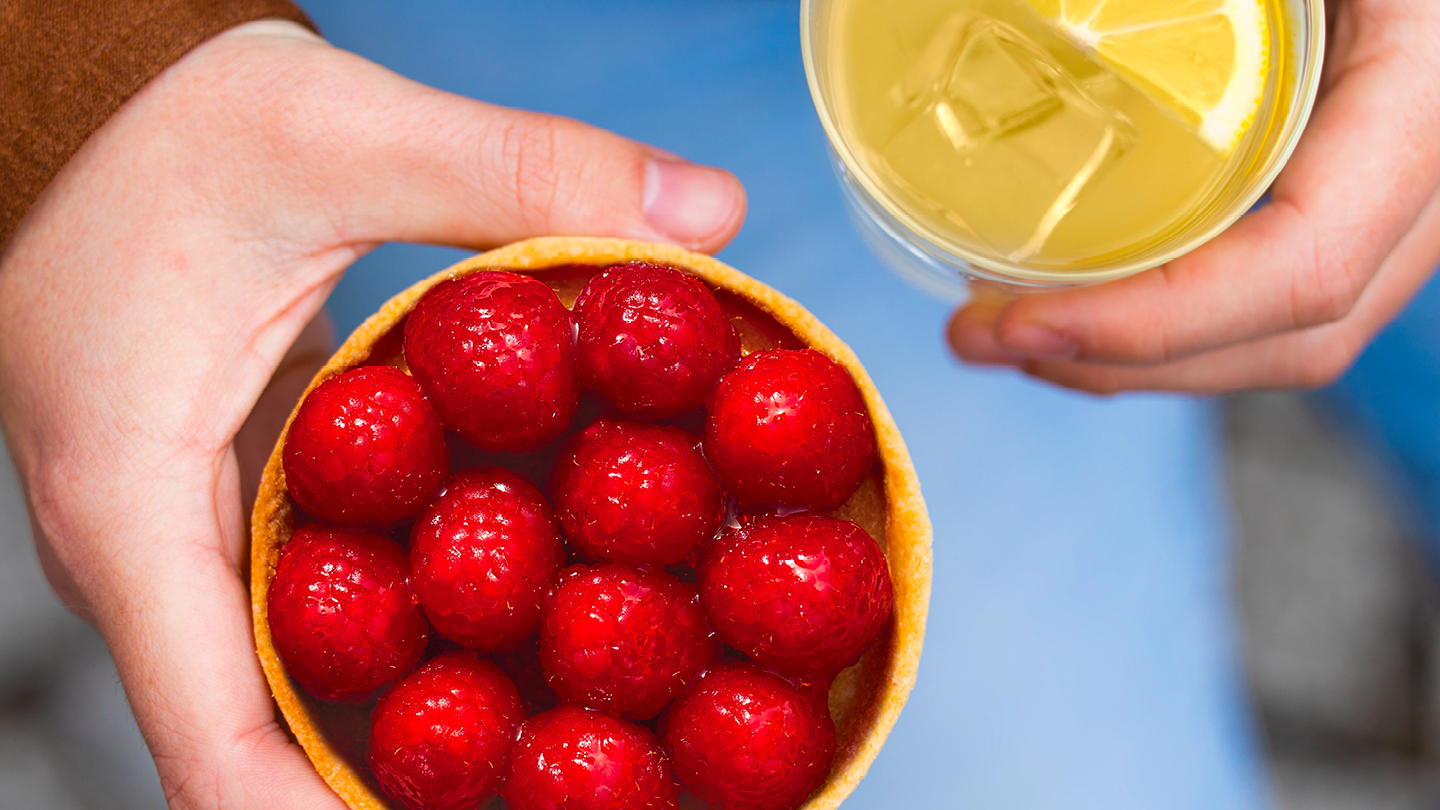 Photo d'une main tenant une tarte à la framboise et un vere de citronnade.